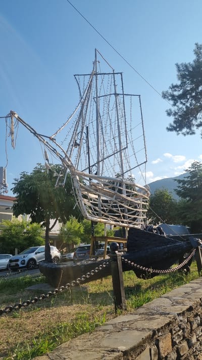 In Litochoro, Greece, an outdoor scene displays a decorative ship structure made of white tubing and string lights, positioned behind a large, black anchor. The ship's frame is elaborate, with numerous lines representing rigging, and is adorned with a dense network of small lights. The anchor is substantial and sits on the ground, secured by a thick metal chain that runs to a post. In the background, trees and a stone wall are visible, along with parked cars and a telecommunications tower. The sky is clear and blue, with a few faint clouds. The overall impression is of a static, decorative display, likely set up for an event or season, situated in an urban or semi-urban environment with natural elements.
