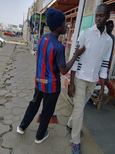 The image depicts a daytime street scene in Mumbwa, Zambia, featuring two adult males in the foreground on an uneven grey interlocking paver sidewalk. The male on the left wears a dark blue beanie, a blue and red striped t-shirt with "UNHCR" text, dark trousers, and light blue and white sandals; his right hand is placed on the left arm of the second male, and his head is turned towards him. The male on the right wears a white collared shirt with black horizontal stripes on the sleeves, beige cargo pants, and dark blue and pink sandals; he is facing the first male. A red and brown broom lies on the pavement between the legs of the male on the left. In the mid-ground, a third male is partially visible behind the rightmost male. The background features several commercial shopfronts on the right, one displaying "ORIFLAME," with merchandise and a red upholstered chair visible. To the left, a dirt road with parked cars, including a red vehicle, and distant pedestrians is visible under bright natural light. A utility pole and a large billboard are also present in the far background.