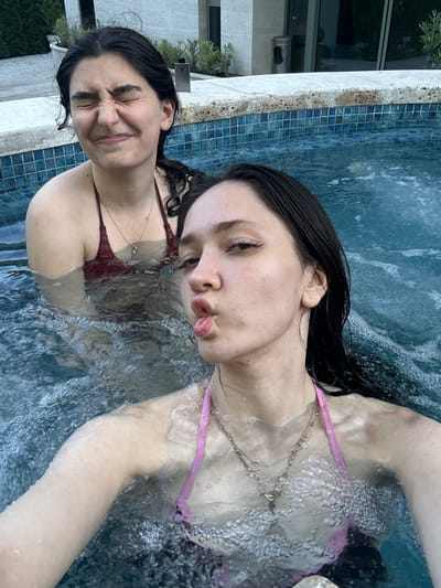 In this media file, two young women are in a swimming pool. The woman in the foreground is closer to the camera and is making a duck face. She has dark hair and is wearing a pink bikini. The woman in the background is slightly further away and is making a silly face with her eyes closed. She has dark hair and is wearing a maroon bikini. The pool water is blue and bubbly, indicating the presence of a jacuzzi or spa. The pool is surrounded by a light-colored tiled edge and a wall with a potted plant. The scene appears to be outdoors, with natural light suggesting it is daytime. The overall mood of the image is playful and relaxed, with both women appearing to enjoy their time in the pool. The location context of Dzoraghbyur, Armenia, does not provide specific visual cues in the image itself. There is no visible text in the image.