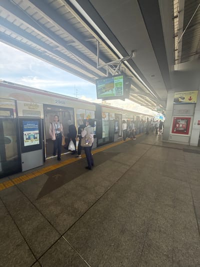 The image depicts a BTS Skytrain station platform in Samrong Nuea Subdistrict Municipality, Thailand. A white train with yellow and orange accents is stopped at the platform, with its doors open. Several people are boarding or disembarking the train. One woman in a pink jacket and dark pants is walking towards the viewer from the train door, holding a phone and a bag. Others are visible in the background, some with shopping bags. The platform is paved with concrete tiles and has yellow tactile paving at the edge. Above the platform, there is a covered walkway with structural beams and a roof. In the background, a digital display shows train information, and various signs are mounted on the station walls. The overall scene is bright, indicating it is daytime, with sunlight filtering through the roof structure.