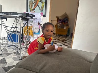 The image depicts a young child indoors in Ilishan, Nigeria. The child, with dark skin and short dark hair, is positioned in the foreground, leaning over the edge of a gray fabric sofa. They are wearing a red short-sleeved top with yellow shoulder accents and are holding a small white bowl with a blue spoon, looking directly towards the viewer.
Behind the child and to the left, a silver-colored, collapsible stand supports DJ equipment, specifically a RANE TWELVE controller and a device labeled "serato RANE." Beneath this stand, a blue hard case with silver latches rests on the floor. Further left and slightly behind the equipment, a white clothing rack is visible, laden with various light-colored garments. On the white wall behind the clothing rack, a framed abstract artwork with colorful, stylized figures is displayed.
To the right and rear, an open doorway with a wooden frame leads into an adjacent room. Through this doorway, a yellow armchair heavily covered with clothing and other items is discernible. The entire visible floor area is covered with black and white geometric patterned tiles. The walls are uniformly white, and the scene is illuminated by ambient light.
