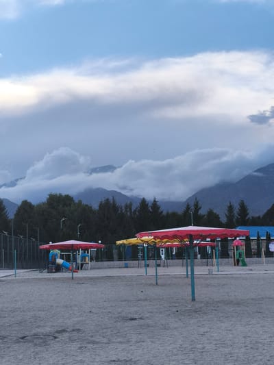 The image depicts an outdoor scene in the city of Bayet, Kyrgyzstan. In the foreground, a sandy area features several brightly colored beach umbrellas, one predominantly red and others with yellow accents. Several children's playground structures are visible behind the umbrellas, including slides and climbing frames. A dark green fence borders the playground area. Beyond the playground, a dense line of dark green coniferous trees forms a backdrop. In the distance, layered mountains rise, with their peaks partially obscured by dramatic, heavy grey clouds. The sky above is a mixture of blue and grey. The overall impression is of a recreational area situated near a natural landscape, under a somewhat overcast sky.