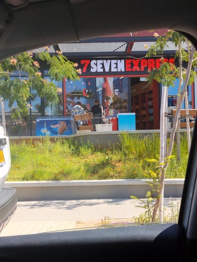 The image shows a view from inside a vehicle looking out at a 7-Eleven Express convenience store in Be'er Sheva, Israel. The store has a prominent red and white "7 SEVEN EXPRESS" sign above its entrance. The store's large glass windows reveal the interior, where two individuals are visible. One person, wearing a patterned shirt and zebra-print pants, appears to be interacting with a display or counter. Another person, in a black top and polka-dot skirt, is also inside. Outside the store, there's a blue refrigerator displaying an ice cream advertisement. A small tree with delicate pink flowers is growing in front of the store, along with lush green grass and some small yellow flowers. The foreground of the image is dominated by the interior of the vehicle, including a portion of the dashboard and windshield frame, with shadows of trees cast on the paved walkway outside.