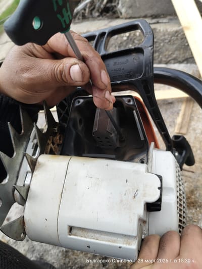 The image depicts a close-up view of a person performing maintenance on a Stihl chainsaw in Balgarsko Slivovo, Bulgaria, on March 28, 2026, at 15:30. A pair of hands, with visible dirt and grime, holds a black and green Wera T-handle screwdriver, inserting its shaft into the exposed internal components of a white and orange Stihl chainsaw. The chainsaw's black handle, marked with "Stihl" and the number "1122 792 91003," is visible at the top. The white housing of the chainsaw shows signs of wear and dirt. A metallic felling dog, exhibiting some rust, is positioned on the left side of the chainsaw. The background consists of a light-colored, possibly concrete or stone, surface, with blurred concrete blocks or a wall and light-colored wooden planks visible further back.