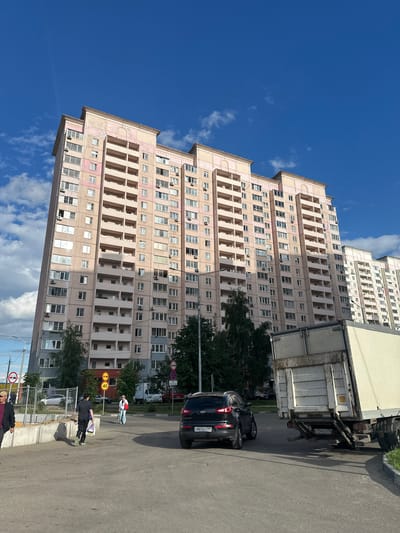 This media file shows a sunny day in Kotelniki, Russia, with a clear blue sky and scattered clouds. The main subject is a tall, multi-story apartment building with a light pink facade. The building has numerous windows and balconies, and air conditioning units are visible on some of them. In the foreground, a gray asphalt road runs alongside the building. A black Kia SUV is parked on the road, and a white delivery truck is also present.
Several people are visible in the scene. One person is walking on the sidewalk, carrying shopping bags. Another person, dressed in a white top and blue pants, is standing near a pedestrian crossing. Two individuals are seen near the sidewalk, and another person in a black jacket is also on the sidewalk. Traffic signs, including speed limit signs indicating 40, are visible on the roadside. The overall scene suggests a typical urban environment on a pleasant day. The time of day appears to be late morning or early afternoon, given the bright sunlight and clear sky. The presence of trees and well-maintained buildings indicates a lived-in residential area.
Visible text includes license plates on the vehicles and text on the traffic signs. The license plate on the Kia reads "O887BT199". The license plate on the truck reads "C124CO799". The speed limit signs show "40".