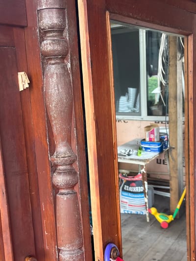 The image shows a close-up view of a weathered wooden door frame with a pane of glass. The door is a dark reddish-brown color and appears to be old, with visible signs of wear and tear, including peeling paint and scratches. A decorative wooden spindle is attached to the door frame on the left side of the image. Through the glass pane, a cluttered outdoor area is visible. A table with various items on it, including containers and a box, can be seen. A large bag, possibly of cement or building material, is leaning against the table. A yellow and green toy duck on a stick is also present on the wooden floor. The background suggests an unfinished or construction area with wooden beams and insulation material. There are no people visible in the image. The scene is likely located in an outdoor or semi-outdoor space, possibly a porch or workshop area.