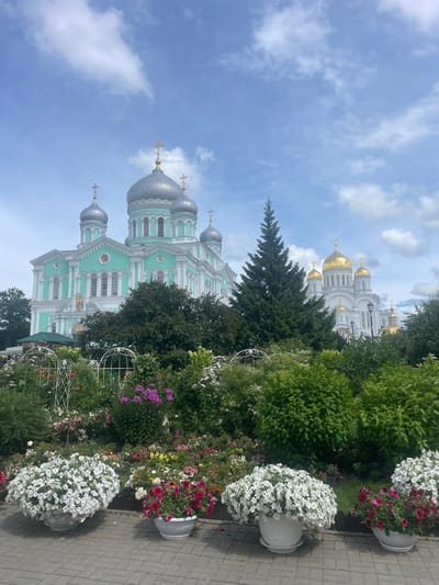 The image displays the Holy Trinity Cathedral in Diveyevo, Russia. The cathedral is a large, ornate building with multiple domes, some silver and some gold. The front of the cathedral is painted a light turquoise color with white architectural details. In the foreground, there is a vibrant flower garden with a variety of blooming plants in various colors, including white, pink, and red. Several large white pots overflowing with white petunias are placed along a paved walkway. A tall, dark green evergreen tree stands to the right of the cathedral, partially obscuring a smaller, white building with golden domes in the background. The sky is blue with scattered white clouds. The scene appears to be a bright, sunny day.
