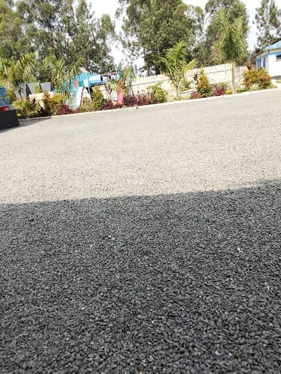 The image depicts an outdoor scene in Malaha Isongo Makunga ward, Kenya, under natural daylight. The foreground and mid-ground consist of a flat, light-grey gravel surface, bisected horizontally by a distinct, dark shadow line in the lower half. Beyond the gravel, a landscaped border features low-lying green and reddish-brown shrubs and several small palm trees. Behind the landscaping, a light-colored wooden fence extends horizontally across the scene. To the left, a children's playground structure, composed of blue, red, and light blue elements, including a slide, is visible. In the background, a dense line of tall, leafy trees dominates the upper portion of the frame, with a segment of a white building featuring blue trim visible on the far right. There are no individuals present.