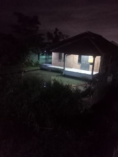 The image captures a quiet, dark night scene featuring a small, single-story house positioned beside a body of water, likely a pond or canal. The house has a dark, tiled roof, brick walls, and a prominent porch or veranda that extends directly over the water. A dark door and at least one window are visible on the house's facade. A light source inside the house or on the porch casts a warm glow, illuminating the structure and creating reflections on the water's surface.
The surrounding area is characterized by dense, dark vegetation and silhouetted trees, suggesting a rural or semi-rural setting. The sky is dark with visible cloud formations, indicating the time of day is night. No people or specific activities are discernible in the static scene. A faint line, possibly a power line, is visible against the dark sky in the upper portion of the image. The overall atmosphere is tranquil and somewhat isolated. No text is visible in the media. This scene aligns with the location context of Kecamatan Pangalengan, Indonesia.