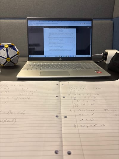 The image shows a desk in an office in Lancaster, UK, at 54.005 latitude and -2.79 longitude. There is a laptop computer on the desk open to a browser window showing a math question. Next to the laptop, on the left-hand side, is a blue and yellow object with 12 sides. Two open notebooks lie on the desk, one on the left and one on the right, with handwritten equations on each. Both notebooks have several black dots across the pages. There is an electrical power adapter on the desk, along with a black bag. The surface of the desk is a dark grey colour, possibly made of wood. There is a grey wall in the background. The lighting in the room is good.