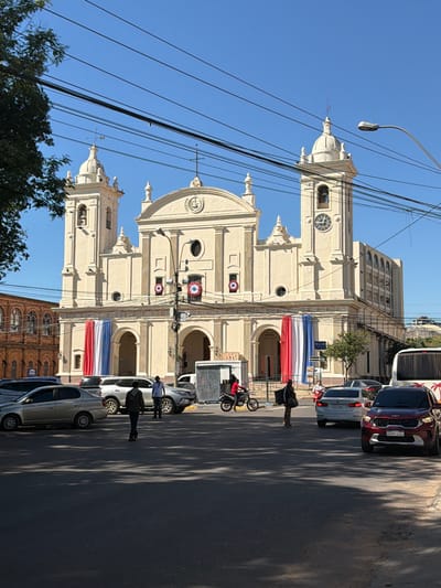 The image depicts a street scene in Asunción, Paraguay, in front of the Metropolitan Cathedral. The cathedral is a large, cream-colored building with twin bell towers and a prominent clock on the right tower. Red, white, and blue flags are draped across the front of the cathedral. The street in the foreground is paved and shows shadows cast by trees. Several vehicles are present, including a red SUV in the foreground with license plate AAOY180, a silver sedan to its left, and a white SUV parked further back. A white bus is visible on the right side of the frame. Two pedestrians are walking in the street, one in dark clothing and another in lighter attire. A person on a motorcycle wearing a red jacket is seen to the left, near the building. Power lines crisscross the sky above the scene. To the left of the cathedral, a building with arched windows, likely a historical structure, is partially visible. The sky is clear and blue.