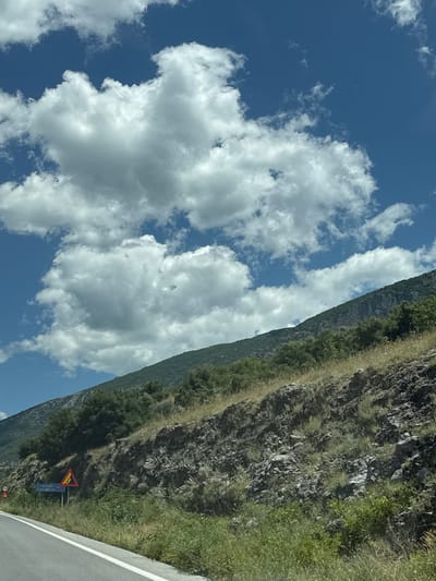 The image captures a section of a road in the Prosotsani Municipal Unit, Greece, under a bright blue sky filled with scattered cumulus clouds. A paved road with a white line marking runs along the left side of the frame. The right side of the road features a steep, rocky embankment covered in dry grass and green vegetation. A forest of trees is visible further up the embankment, merging with the dark green slopes of a mountain range that dominates the background. Two road signs are present: a triangular yellow sign with a black symbol indicating a dangerous curve or turn, and a blue rectangular sign with white lettering, partially obscured by vegetation, likely indicating a place name or direction. In the far left of the image, a circular red and white sign with the number "70" is visible, suggesting a speed limit. The overall scene depicts a rural or mountainous area, likely along a roadway.