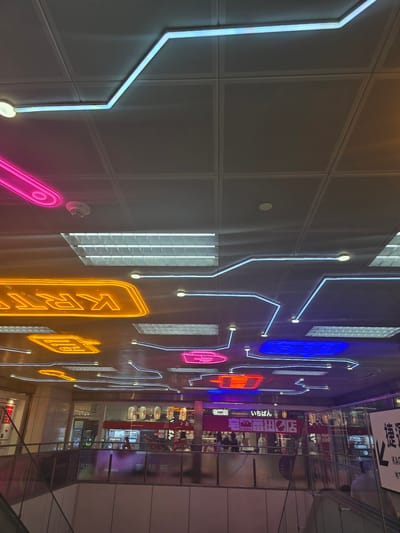 This image captures a low-angle view of a ceiling adorned with a network of blue and white LED strip lights, creating a futuristic, circuit-board-like pattern. Below, neon signs in vibrant pink, orange, and blue cast a colorful glow on the reflective ceiling panels. Fluorescent light fixtures are also visible.

The scene appears to be in an indoor public space, possibly a shopping mall or an entertainment complex, given the presence of escalators and store facades visible in the background. Store signage includes Japanese characters, such as "一番" (Ichiban) and "宅麺" (Taku-men), suggesting a connection to Japanese cuisine or culture. One sign also has Chinese characters, "優惠券" (Yōu Huì Quàn), meaning "coupon."

The location is identified as Kaohsiung, Taiwan. Specifically, this appears to be the interior of the Kaohsiung MRT Station, likely near the entrance to the underground shopping street, which features themed neon lighting and has a notable Japanese influence. There are several individuals visible in the background, some walking and others appearing to be patrons of the shops. The overall atmosphere is modern and energetic, illuminated by a combination of artificial lights.