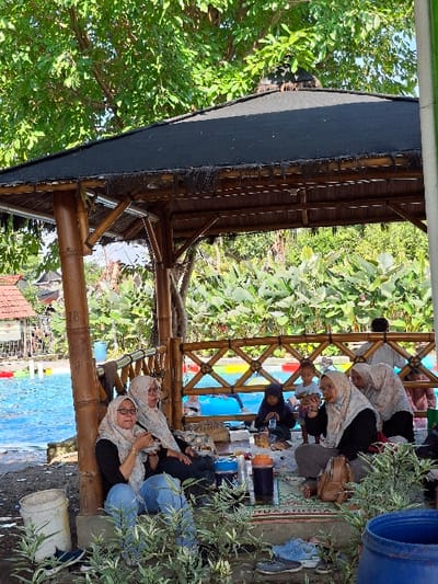 The image shows a group of people sitting under a bamboo hut with a black tarp roof at a swimming pool in Kecamatan Tulung, Indonesia (coordinates: -7.592, 110.65). The group is composed of five women and one young boy. Two of the women are wearing headscarves and are sitting at the table, engaging in conversation and eating from bowls. The remaining three women are sitting on the ground nearby. The young boy is wearing a light blue shirt and shorts. The setting is outdoors and the scene is sunny and warm. The environment consists of a swimming pool, green grass, dense foliage, a large bamboo structure, and a blue plastic bucket.
