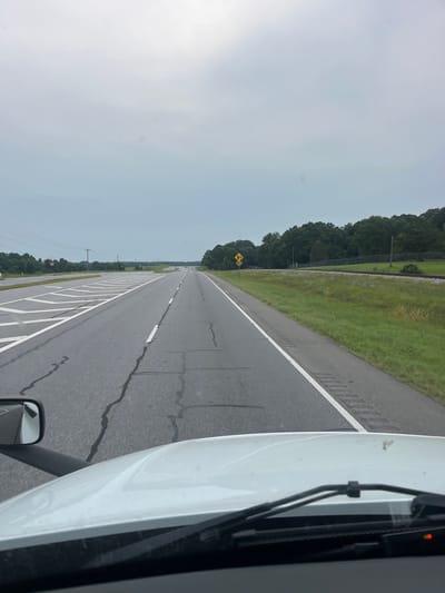 The image captures a view from the cab of a white truck, looking forward on a two-lane highway. The road is asphalt with visible cracks and white lane markings. On the left side of the road, there are complex white markings indicating a merging or diverging lane, and further in the distance, power lines and trees can be seen. To the right of the road, there is a grassy area with a fence and a railroad track running parallel to the highway. A yellow road sign with a black symbol indicating a sharp curve is visible on the right shoulder. The sky is overcast and gray, suggesting a cloudy day. The foreground shows the windshield, wipers, and a side mirror of the truck. The scene appears to be in a rural or suburban setting.