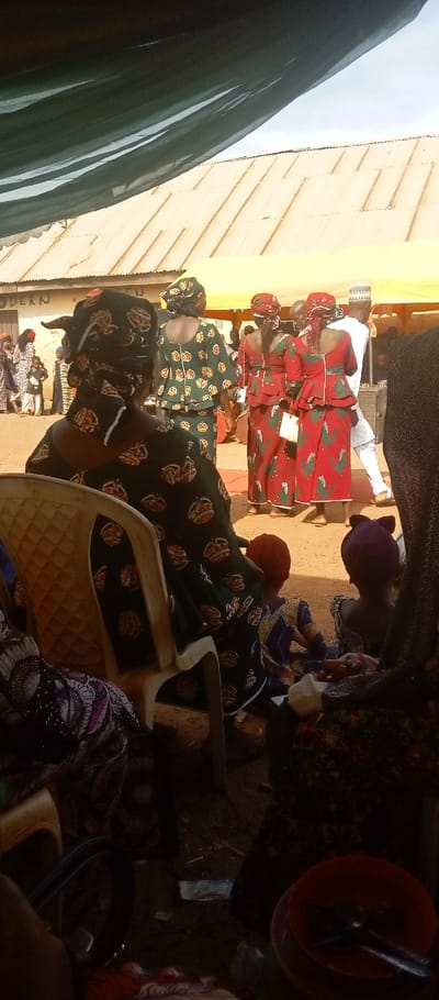 An outdoor gathering is depicted in Kasuwan Magani, Nigeria, viewed from under a dark canopy. The ground consists of dry, reddish-brown earth. In the foreground, a person is seated on a light-colored chair with their back to the viewer, wearing a dark green patterned top, skirt, and headwrap. To the right, two young children are visible from behind, seated on the ground; one wears a red head covering and a blue/gold patterned outfit, the other a purple head covering. In the lower right corner, an orange bowl containing two spoons rests on the ground.
In the midground, multiple individuals are standing. Three women are visible from behind: one in a green patterned outfit and headwrap, and two in matching red patterned outfits and red headwraps, one holding a small white bag. To their right, a man in a white tunic and white cap is partially visible, facing away. A large drum is present on the ground. A yellow canopy structure extends across the midground.
The background features a building with a light-colored corrugated metal roof. A partial sign on the building wall reads "MODERN." A group of people, including adults and children, are visible in the distant background near this building. The sky above is pale blue.