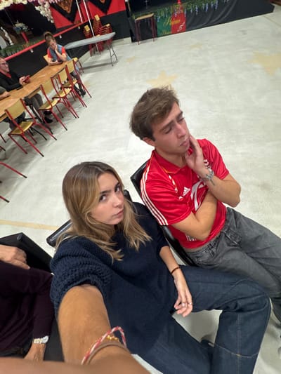 The image displays an indoor event space located in Décines-Charpieu, France. In the foreground, a light-skinned female with shoulder-length blonde hair, wearing a dark blue knitted sweater and dark blue jeans, is seated, holding a recording device with her extended right arm. She wears a multi-colored bracelet on her right wrist, a reddish-brown bracelet on her left, and a ring on her left hand, looking directly at the camera. To her immediate left, a partial view of another individual shows a dark purplish long-sleeved shirt and a gold-toned wristwatch. To her right, a light-skinned male with short brown hair, wearing a red short-sleeved polo shirt with white shoulder stripes and dark grey jeans, is seated. He wears a silver-toned bracelet and a tattoo on his left forearm. His right hand contacts his right cheek/jawline as he looks downwards with a pensive expression.
The background features a light-colored floor adorned with large, faded yellow star patterns. Multiple light-colored wooden tables with red-framed chairs are arranged in rows. Two additional individuals are visible seated at one of these tables on the left: a light-skinned male in a dark shirt, and a light-skinned female with hair pulled back, wearing a light blue sleeveless top over a red shirt, leaning forward. The rear wall is dark, featuring red and black fabric drapery, white artificial floral decorations, and a banner displaying red, green, and white stripes with the text "Paião," "Dança," and the numeral "5." Stacks of red-framed chairs and additional folding tables are visible along this wall.