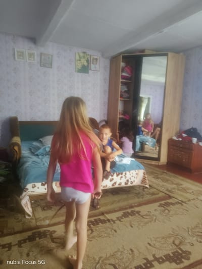 This media file depicts a candid indoor scene, likely a bedroom in a home, given the presence of a bed, a wardrobe with a mirrored door, and decorative wallpaper. The setting appears to be in Sestrynivka, Ukraine. Three children are visible. In the foreground, a young girl with long blonde hair, wearing a bright pink t-shirt and white underwear, is walking away from the camera towards a bed. She is barefoot. On the bed, a young boy wearing a blue t-shirt is seated and looking towards the camera. Behind him, another child, possibly a girl, wearing a purple and white striped top, is partially visible. The mirrored wardrobe reflects another child, a girl in a pink top, standing in the reflected room. The overall atmosphere seems relaxed and informal, suggesting a moment of everyday life. The lighting suggests it is daytime. The wallpaper features a repeating floral pattern, and several framed pictures are mounted on the wall. A patterned rug covers the floor. The bottom left of the image displays the text "nubia Focus 5G".