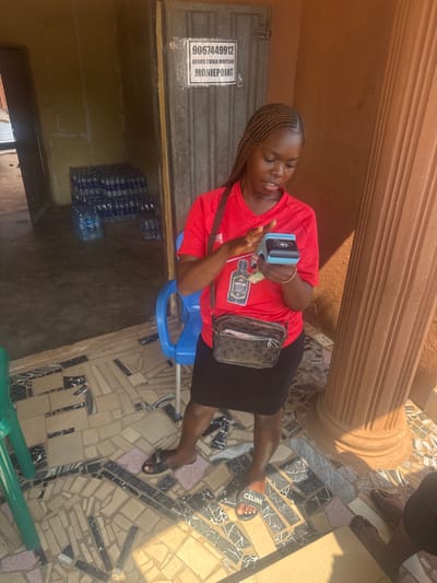 The image depicts a young adult female standing on an irregularly tiled outdoor or semi-outdoor surface in Aguata, Nigeria. The subject has dark skin and braided hair, wearing a red short-sleeved t-shirt with a dark bottle logo, a black skirt, and black "Celine" branded slide sandals. A dark brown patterned crossbody bag is visible on her left side, and a gold-colored bracelet is on her right wrist. She holds a light blue and black portable point-of-sale (POS) device in her left hand and is operating its screen with her right index finger. Nigerian Naira banknotes are partially visible in her left hand beneath the device.

In the background, a metallic door features a white sign displaying "9067449912", "OKORO CHIKA MARYANN", and "MONIEPOINT". To the left of the door, an open doorway reveals an interior area with pale yellow walls, where multiple stacks of shrink-wrapped plastic water bottles are visible on the floor. A blue plastic chair is partially obscured behind the subject. To the subject's right, a fluted brown pillar is prominent, casting a shadow due to strong natural sunlight. The ground surface is composed of a mosaic of light, dark brown, and black tile fragments. The lower right foreground shows the partial view of another individual's legs and feet, wearing brown sandals.