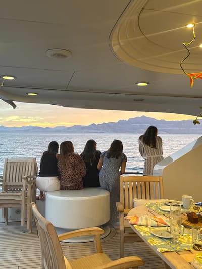 Captured from the perspective of a yacht's interior, the image frames a group of individuals observing the coastline of an unknown location in Mexico. The foreground showcases a dining table with tableware set for a meal. A semi-circular seating area accommodates multiple individuals, identifiable by their varying clothing patterns and hairstyles. The background reveals a mountainous coastline meeting the ocean, with hues of sunlight suggesting either sunrise or sunset. Interior yacht features include ceiling lights, curved wall designs, and deck flooring.