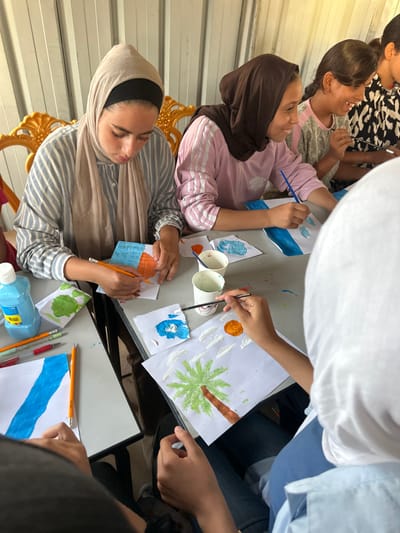 The image depicts an indoor art activity involving five young females in a-Zawayda, Palestinian Territory. The scene features light-colored corrugated metal walls in the background and two ornate gold-colored chairs. Three grey tables are arranged, laden with art supplies.

In the immediate foreground, a female wearing a white hijab and a light blue top uses a paintbrush to add white textured elements, possibly clouds, to a drawing on white paper. Her artwork includes a green palm tree with a brown trunk and an orange sun. To her left, another female, dressed in a beige hijab and a grey-and-white striped long-sleeved shirt, focuses intently on her paper, using a yellow pencil to color an orange shape next to a blue rectangle. On the table between these two individuals are two white paper cups, one containing blue paint residue and another with green. A blue plastic bottle and several colored pencils are also visible on the table.

Further into the scene, a third female, wearing a brown hijab and a pink striped long-sleeved shirt, uses a paintbrush to apply blue stripes to her white paper. To her right, a fourth female with dark hair and a patterned shirt smiles while also painting. A fifth female is partially visible further to the right. Each participant is engaged with their own individual paper and art supplies, working on various drawings and paintings.
