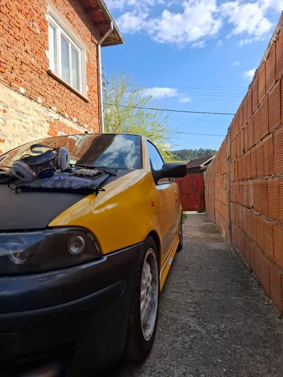 A yellow vehicle is parked tightly between two brick buildings in Rakitovo, Bulgaria. The sky is visible above, with some white clouds. The building on the left has a window. The vehicle has a black front bumper and safety gear attached to the hood.