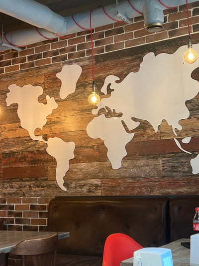 The image shows a close-up of a wall decorated with a white outline of the world map on a wooden plank background. Above the map, exposed grey pipes with red wiring are visible against a brick wall. Several Edison-style light bulbs hang down, illuminating the scene. In the foreground, a dark brown leather booth and a glimpse of a table and chairs are visible, suggesting an indoor seating area, possibly a restaurant or cafe. The lower right corner shows a bottle of Coca-Cola on a table. The scene does not provide any specific visual cues to determine its location in Mineralnye Vody, Russia.