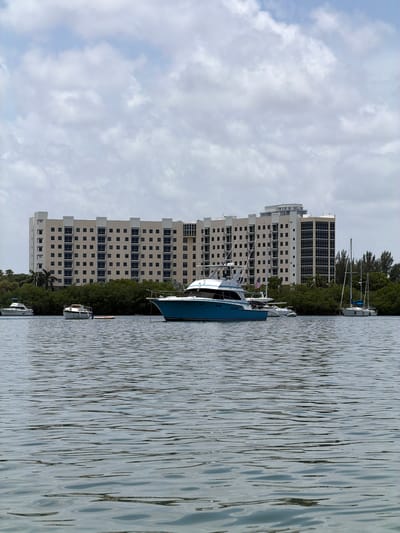 A wide shot captures a body of water in the foreground with a large, multi-story building and a line of trees in the background. The water is choppy, with ripples and reflections creating a textured surface. Several boats are docked or anchored in the water. The closest boat, a large blue and white yacht, is positioned in the lower right quadrant of the image. To its left, a smaller white boat with a blue trim is anchored. Further left, another white boat is visible, and a small dark object is floating in the water between the two smaller boats. In the background, a dense line of green trees forms a natural barrier, behind which stands a long, light-colored building with numerous windows. The building appears to be a condominium or apartment complex. The sky above is overcast with patches of blue visible through the clouds. This scene is located in North Miami, Florida, United States of America, likely along the Intracoastal Waterway or a similar canal.