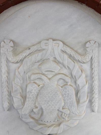 A white stone relief carving, likely marble, is prominently displayed in a vertical orientation. The central subject is a double-headed eagle, depicted with scaled feathers, spread wings, and two crowned heads. This eagle is encircled by a symmetrical wreath composed of stylized foliage. Above the wreath, a horizontal braided band extends, terminating at each end with a four-petaled floral rosette. The carving is integrated into a smooth, light-grey stone surface, with a curved segment of reddish-brown brickwork visible along the upper edge. Minor particulate accumulation is present within the carved recesses and on the flat stone surface. This architectural element is located in Gigintsi, Bulgaria.