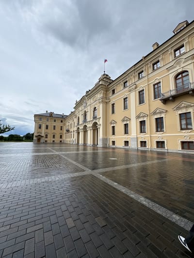 The image shows a wet, paved courtyard in front of a large, pale yellow, classical-style building, likely a palace or government building, located in Strelna, Russia. The sky is overcast with gray clouds. The building features multiple stories with numerous windows, arched doorways, and ornate architectural details. A Russian flag flies from a flagpole on the roof. The courtyard pavement is made of dark rectangular bricks arranged in a patterned, and it is wet, reflecting the sky and the building. In the lower right corner, the lower legs and feet of an individual wearing black pants and white-and-black sneakers are visible. The scene appears to be captured during the daytime.