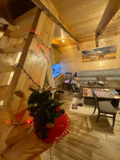 An interior view of a room in the city of Palež, Montenegro, is depicted, characterized by extensive light-toned wood paneling on the walls and ceiling, featuring exposed wooden beams. In the left foreground, a substantial wooden staircase structure, adorned with red LED string lights, rises diagonally. Positioned on a surface in front of the staircase is a small, artificial tabletop Christmas tree decorated with red ornaments and pinecones, resting on a red, scalloped placemat with a snowflake pattern. A dark object is partially visible beneath the tree.
To the right, a large, L-shaped grey sectional sofa with brown cushions is visible. In front of the sofa are two dark-colored low tables. The table closer to the sofa displays several board game boxes, including one labeled "Gloomhaven." The table further forward has a board game fully set up and in progress, featuring numerous game components. A dark grey upholstered chair is positioned at this table. To the left of the sofa, a single dark wooden chair is present, with a pink object on its seat, and a woven basket rests on the floor nearby.
On the back wall, a rectangular painting depicting two horses grazing in a field with snow-capped mountains hangs above the sofa. In the left background, a television screen, mounted on a light-colored brick structure, displays a scene of multiple figures on a snowy landscape, possibly engaged in winter sports. The room is illuminated by warm overhead lighting from an exposed bulb and other fixtures. The floor is covered with light-colored plank flooring. No individuals are visible in the scene.