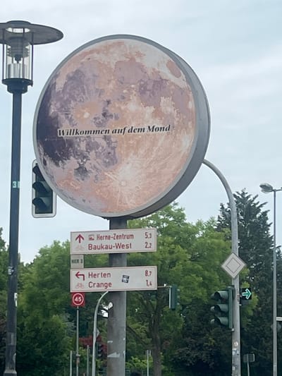 The image shows a large, circular sign depicting the moon with the text "Willkommen auf dem Mond" (Welcome to the Moon) superimposed on it. Below this sign, there are several directional street signs. One sign points upwards and to the right, indicating "Herne-Zentrum" (Herne Center) and "Baukau-West" with corresponding distances of 5.3 and 2.2 respectively. Another sign points to the left, indicating "Herten" and "Crange" with distances of 8.7 and 1.4. To the left of the directional signs, a traffic light is visible. Further down the pole, a speed limit sign with the number "45" inside a red circle is present. The environment appears to be an urban setting with trees and a cloudy sky. The presence of the directional sign for "Herne-Zentrum" confirms the location in Herne, Germany.