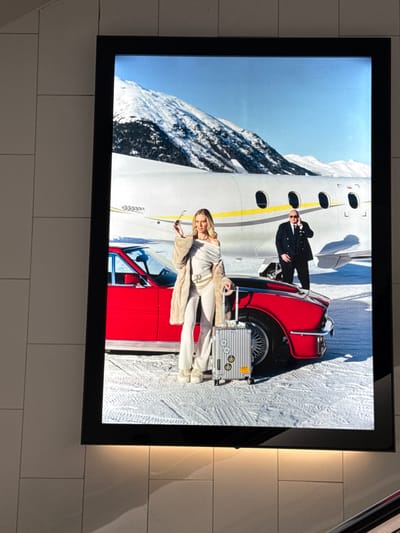 The image shows a large, illuminated advertisement displayed in what appears to be a public space in Sesto San Giovanni, Italy, possibly a shopping mall or transportation hub, given the tiled wall. The advertisement depicts a glamorous scene set against a backdrop of snow-covered mountains and a private jet. A woman in a white off-the-shoulder top and matching pants, accessorized with a fur coat and a belt bag, stands next to a bright red vintage car. She holds a pair of sunglasses in her hand. Beside her is a silver suitcase with stickers. A man in a dark suit and tie stands behind the car, appearing to be on a mobile phone. The ground is covered in snow, with tire tracks visible. The advertisement is framed by a black border and is lit from behind.