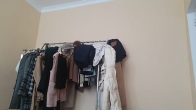A clothing rack filled with various garments dominates the view. Coats in shades of gray, beige, black, and pink hang along the rack. A white puffer coat is also present, along with a blue jacket. The rack is positioned against a light beige wall. A portion of the room's ceiling, trimmed with white molding, is visible at the top of the frame.