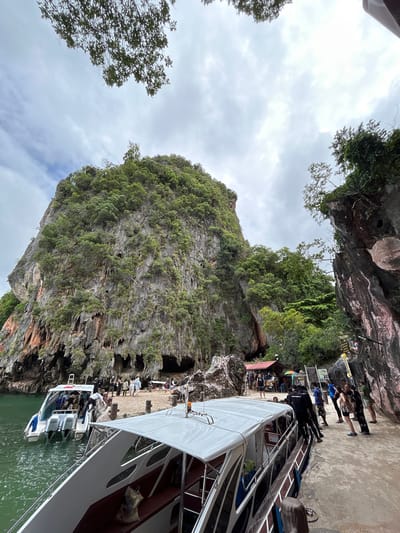 In Tambon Krasom, Thailand, a dramatic limestone karst cliff rises from emerald green water. Several speedboats are docked, with passengers disembarking and boarding. People are gathered on a concrete walkway that hugs the base of the cliff, some looking towards the water and boats, while others are milling around small shops and stalls set up against the rock face. The sky is overcast, with scattered clouds. The scene depicts a popular tourist destination, likely a departure point for island tours.