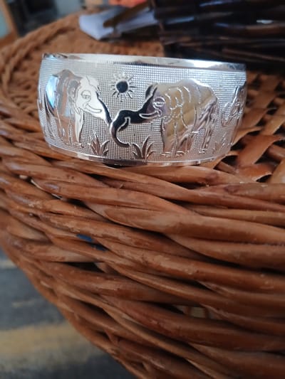 This is a close-up, eye-level, outdoor shot of a silver bracelet featuring an embossed elephant design. The bracelet is resting on a woven brown surface, possibly a basket or placemat. In the background, out of focus, are some dark, elongated objects and a white surface with some writing on it, suggesting an indoor environment or a table setting. The lighting is natural, casting reflections on the polished silver of the bracelet. There are no people present in the image, and no specific landmarks of Póvoa de Varzim, Portugal, are visible. The focus is entirely on the bracelet and its immediate surroundings.