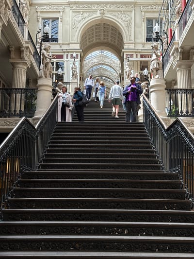 The image shows a grand, ornate staircase inside a covered passage. The staircase is made of dark metal with intricate carvings on each riser, and it ascends towards a brightly lit upper level. Several people are visible on the stairs, walking in both directions. On the left side, a woman in a long light-colored coat is descending, while a woman in a dark jacket is also descending on the right. Further up the stairs, a group of people are walking upwards, including a man in a purple jacket, a man in a white sweatshirt, and two women. The architectural style suggests a historic building, likely a shopping arcade or a grand public building. This scene is located within the Passage Pommeraye in Nantes, France, a renowned 19th-century shopping arcade.