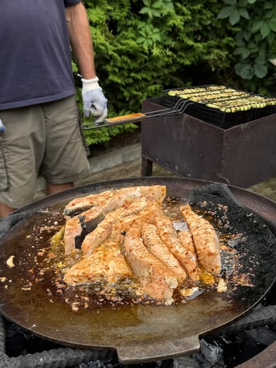 In Rezekne, Latvia, an outdoor cooking scene unfolds with a focus on grilled fish. The primary subject is a large, round metal pan filled with several pieces of seasoned fish, likely salmon given their color and shape, cooking over hot coals. The pan is placed within a dark, round grill. To the left of the grill, a person's arm, wearing a blue garment and a white and blue checkered glove, is visible, holding a grill spatula with a wooden handle. This person appears to be tending to the cooking fish. Behind the person and the grill, a lush green hedge and a metal grill rack filled with what appears to be vegetables or more fish are visible. The scene suggests a casual outdoor meal preparation, likely during daylight hours given the lighting. The overall impression is one of preparing a delicious meal in a relaxed, natural setting. There is no visible text in the image.