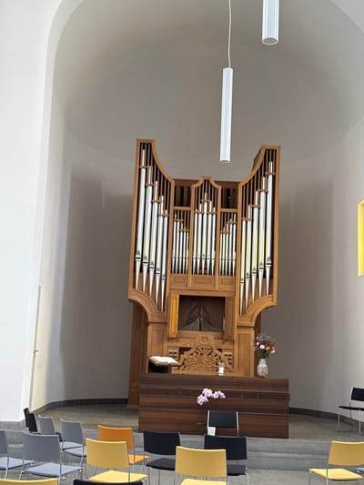 The image depicts the interior of a modern church sanctuary, likely in Sesto San Giovanni, Italy, characterized by its minimalist design and architectural features. Dominating the scene is a large, wooden pipe organ with gleaming silver pipes arranged in a decorative manner. The organ is situated on a raised platform behind a wooden altar or communion table. On the altar, a book is open, and a vase of flowers, along with a glass of water, are present. The wall behind the organ features a subtle architectural curve, and two cylindrical fluorescent light fixtures hang from the ceiling.
In the foreground, rows of chairs are arranged on a tiered floor. The chairs vary in color, with some being grey, yellow, orange, and black. The floor is tiled in a neutral tone. The overall lighting is soft, casting gentle shadows. The environment appears to be clean and well-maintained, suggesting an active place of worship. The architectural style is contemporary, with smooth, curved surfaces and clean lines. The limited number of visible people, primarily implied by the presence of the chairs, suggests a moment before or after a service.