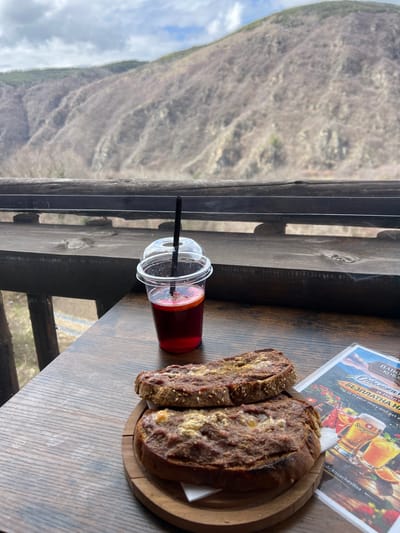 The scene is a view from an establishment in Kovachevitsa, Bulgaria. A wooden table, likely on a deck, features a plastic cup of red drink with a black straw, a wooden platter holding two slices of bread, and a menu featuring a beer advertisement with the word "Staroprame" visible. The view from the deck looks out over a mountainous landscape with sparse vegetation. The sky is overcast with clouds.