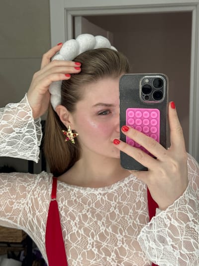 A woman takes a selfie in a mirror. She is fair-skinned with light brown hair pulled back, featuring a white padded headband. A star-shaped earring with red and green gems is visible. She wears a white lace top with red straps. Her nails are painted red. The phone is dark-colored with a pink bubble-pop grip attached. The background includes a window and indistinct furnishings.