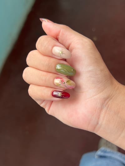 The image shows a close-up of a person's left hand with an oval-shaped manicure. The nails are painted in varying colors and designs. The thumb nail is a light beige with a gold starburst accent. The index finger is painted a muted olive green with a gold curved line and two small gold dots. The middle finger has a marbled design with shades of pink, red, and gold accents, including a gold leaf-like element. The ring finger is a deep burgundy red with a gold starburst accent. The fingernails are all well-maintained and appear to be acrylic or gel extensions. The skin on the hand is light-toned. The background is blurred, with a dark brown, wooden surface visible below and a lighter teal-colored surface to the left. The lighting suggests an outdoor setting. There is no discernible location in the city of Villa de Cura, Venezuela depicted in this image.