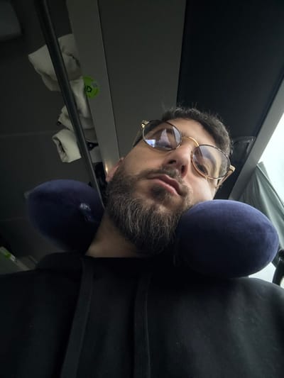 A low-angle, close-up photograph captures an adult male resting inside a public transportation vehicle, likely a bus or train, in Rosny-sur-Seine, France. The subject, positioned centrally, has a short dark beard with grey highlights and short dark hair. He wears gold-framed glasses with a prominent upper brow line, reflecting light. His eyes appear heavy-lidded or closed, suggesting sleep or rest. A dark blue, U-shaped travel neck pillow supports his head. He is dressed in a black hooded sweatshirt with visible drawstrings. In the upper left background, an overhead storage compartment contains several folded white fabric items and a small green circular sticker with a white logo. A muted, diffused light source is visible through a window on the upper right, indicative of an overcast sky or interior lighting.
