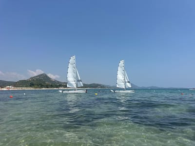 Two small sailboats with white triangular sails are floating on a clear blue sea under a bright sky. The sailboats are identical in design, with white hulls and black accents on their sails. They are positioned in the middle ground of the image, with the one on the left slightly closer to the viewer. In the background, a sandy beach is visible, lined with rows of white umbrellas and some beach structures. Beyond the beach, a green, hilly landscape rises, with a prominent peak in the center. The water in the foreground is very clear, allowing the sandy bottom to be seen. Several buoys, one red and some white, are scattered in the water. Two people can be seen swimming or wading in the sea between the sailboats. The scene appears to be a sunny day at a beach resort in the municipality of Castiadas, Sardinia, Italy.