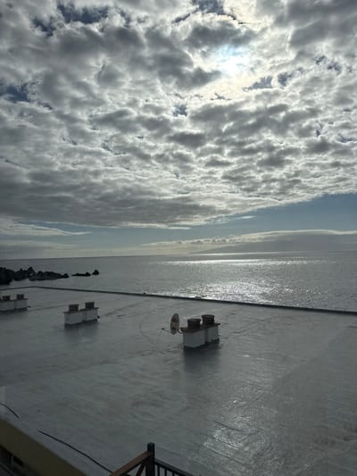 The image captures a panoramic view of the Atlantic Ocean and a flat rooftop from an elevated position in Santiago del Teide, Spain. In the foreground, a light grey or off-white rooftop, possibly covered with a waterproof membrane, is visible. It features three rectangular utility structures, two grouped on the left and one isolated on the right, each topped with two dark, cylindrical vents. A small, white, circular satellite dish is affixed to the rightmost utility structure, with connecting wires visible on the rooftop surface. A dark railing, likely made of wood or metal, is partially visible in the lower left corner, indicating the viewpoint.
Beyond the rooftop, the expansive ocean extends to the horizon. On the left side, dark, rugged volcanic rock formations are present along the coastline, with waves breaking around them. Across the water, in the mid-ground on the right, a distant, hazy landmass with undulating terrain is discernible. The sky is largely covered with an uneven layer of altocumulus clouds, exhibiting varied texture and density. The sun is partially obscured by clouds in the upper right portion of the sky, casting a strong, bright reflection across the ocean's surface, creating a path of glinting light extending towards the viewer. Patches of blue sky are visible between the clouds. No people or animals are present in the scene.