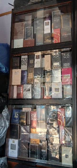 A vertical image depicts a wooden retail display cabinet with glass doors, stocked floor-to-ceiling with various boxed perfumes in Sabon Gari South, Nigeria. The cabinet features at least three visible shelves, each densely packed with fragrance products, most of which are still sealed in cellophane wrapping. Visible brands include "212 MEN NYC", "212 SEXY MEN", "SAUVAGE", "DIOR", "TERRE D'HERMÈS", "PURE XS", "WANTED", "GIORGIO ARMANI SI", "AZZARO", "CHANEL", "MONT BLANC", "LACOSTE", "GUCCI", and "JO MALONE". The immediate environment outside the cabinet on the left side shows dark, indistinct shapes, possibly bundled materials or merchandise. No individuals are present within the frame. The scene represents a static inventory display within a commercial establishment.