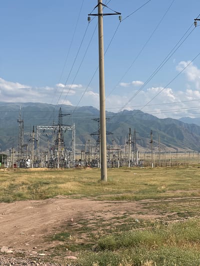 This media file depicts a power substation set against a backdrop of mountainous terrain and a clear blue sky with scattered clouds. The substation is a complex arrangement of towers, transformers, and electrical lines, suggesting an active electrical infrastructure. A large concrete pole and numerous power lines dominate the foreground, extending across the frame and connecting to the substation in the midground.
The scene is set in a rural or semi-rural area, indicated by the presence of dry, grassy fields and a dirt path running alongside the substation. The vegetation appears sun-baked, and the overall lighting suggests it is daytime, likely a sunny afternoon. The distant mountains are layered and appear green and brown, hinting at the geographical location in Kazakhstan, specifically within the Sarymoldayev rural district.
There are no people visible in the frame, and the overall mood is one of industrial infrastructure in a natural landscape. The focus is on the electrical equipment and its integration into the environment. The image is taken from a slightly low angle, emphasizing the height of the power lines and the scale of the substation. No visible text is present in the image.