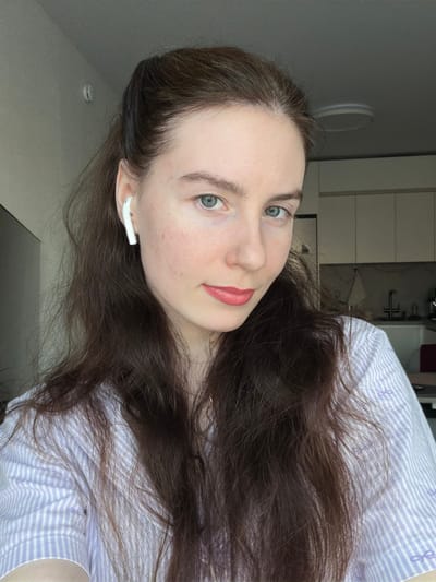 A female individual is depicted from the chest up in an indoor residential setting. She has fair skin, blue-green eyes, and dark brown wavy hair, partially pulled back from her face, with strands falling over her shoulders. She wears pinkish-red lipstick and a light purple and white striped shirt adorned with small purple bow patterns. A white Apple AirPods earbud is visible in her right ear. Her gaze is directed towards the camera. The background partially shows a modern kitchen area on the right, featuring white upper cabinets, a sink with a faucet, and a light-colored wall. A round, flush-mount light fixture is visible on the white ceiling. A red object is partially visible on a counter in the background. The specific location within the city of Ковалёво, Russia, is not discernible from the image content.