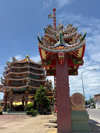 The image shows the exterior of a colorful Chinese temple in Ang Sila, Thailand, under a bright blue sky with scattered clouds. The temple features ornate multi-tiered roofs with intricate dragon and floral decorations, painted in vibrant reds, yellows, blues, and greens. One of the main structures in the foreground is a tall, reddish-brown pillar supporting a decorative roof section. To the left, a multi-layered hexagonal building is visible, adorned with detailed murals depicting scenes and figures. In front of this building, there are trees and shrubs. A street with traffic is visible in the background, suggesting the temple is located in a developed area. A street lamp is also present to the right of the pillar. Near the pillar, on a green base, is a circular monument with a Chinese character inscribed in its center. The ground in the foreground is a mixture of dirt and paved areas.