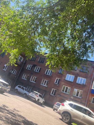 In Kiselyovsk, Russia, on a sunny day, a multi-story brick apartment building stands beneath a canopy of lush green leaves from an overhanging tree. The building's facade features numerous windows, many with white frames, interspersed with a few dark-framed windows and some with a blue tint. A narrow balcony juts out from one of the lower floors.
In front of the building, a paved area serves as a parking lot for several vehicles. A white SUV, a white compact car, and a darker colored car are visible. The white SUV, prominently positioned in the foreground, is angled towards the right of the frame. The ground reflects the shadows of the trees, creating a dappled pattern. The sky is a clear blue.